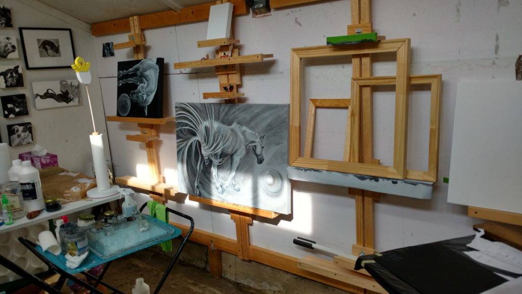 a wall of wooden easels and two black and white paintings of horses with balls, among other canvases, disrupted by rectangles of light cast from a window